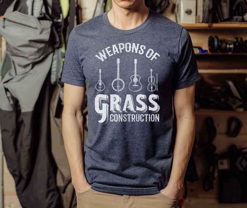 Op de afbeelding: Een marineblauw t-shirt met een wit grafisch ontwerp met de tekst "Weapons of Grass Construction" en een rij van vijf muziekinstrumenten, waaronder een banjo, een gitaar en een viool.