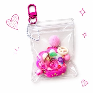 May include: A clear plastic pouch with a pink lobster clasp and a ball chain. Inside the pouch is a pink, white, and purple charm featuring two glittery red cherries, a small pink pom-pom, a white peace sign, and various colorful beads.