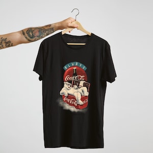 May include: Black t-shirt featuring a Coca-Cola design. The graphic shows a polar bear wearing sunglasses, relaxing on a Coca-Cola can, with the words "Always Coca-Cola".