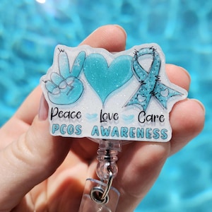 May include: A white glitter badge holder with a peace sign, heart, and blue ribbon. The text "Peace Love Care PCOS AWARENESS" is printed on the badge holder.