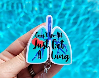 Op de afbeelding: Een witte plastic badgehouder in de vorm van een long met de tekst "Can't We All Just Get A Lung" in zwart. De long is blauw en heeft een rood hart erin.