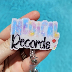 May include: A white badge holder with a glittery rainbow design and the text "Medical Records" with a red plus sign. The badge holder is attached to a clear retractable badge reel.