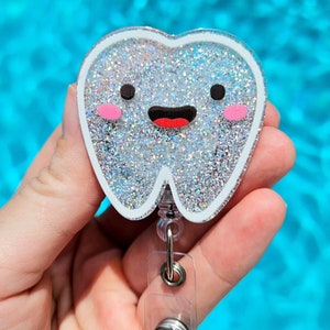 May include: A silver glitter tooth charm with a smiling face and pink cheeks attached to a clear retractable badge holder.