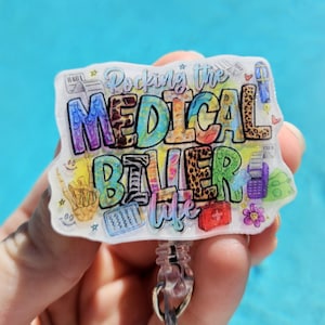 May include: A white badge holder with a colorful design that says "Rocking the Medical Biller Life". The design includes a stethoscope, a medical chart, and a first aid kit. The badge holder has a clear plastic retractable reel.