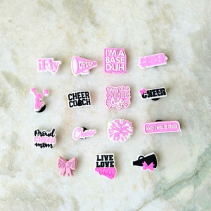 May include: A collection of pink and black cheerleading shoe charms with various sayings, including "I'm a base duh", "Cheer Coach", "Live Love Cheer", and "Sorry I was at cheer".