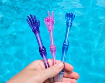May include: Three colorful plastic pens shaped like hands. The pens are blue, pink, and purple with a gradient effect. The pens are held in a hand against a blue pool background.