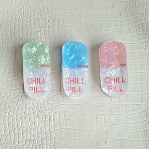 May include: Three pill-shaped decorative items with a textured, semi-translucent finish. Each features a colored top half in light green, blue, or pink, and a white bottom half. The text "CHILL PILL" is printed in red, with "100mg", "200mg", or "15mg".