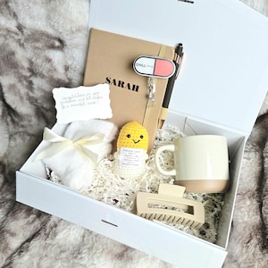 May include: A white gift box containing a tan notebook personalized with the name SARAH, a black pen, a pill-shaped charm labeled CHILL, a white card with handwritten text, a white pair of socks with a ribbon, a yellow crochet doll, and a mug.