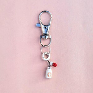 May include: A silver-colored keychain with a lobster clasp, a small ring, and a charm. The charm is a white pill bottle with a red pill on top. The pill bottle has a gold cross design.