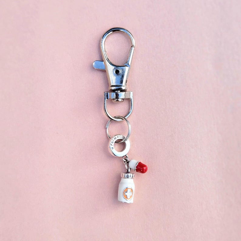May include: A silver-colored keychain with a lobster clasp, a small ring, and a charm. The charm is a white pill bottle with a red pill on top. The pill bottle has a gold cross design.