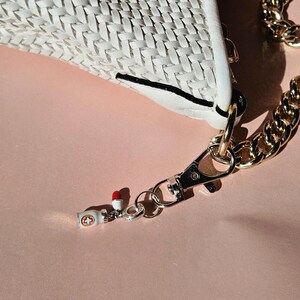 May include: Close-up of a white woven handbag with a gold chain strap. A small charm with a red and white pill design hangs from the clasp. The background is a soft pink.