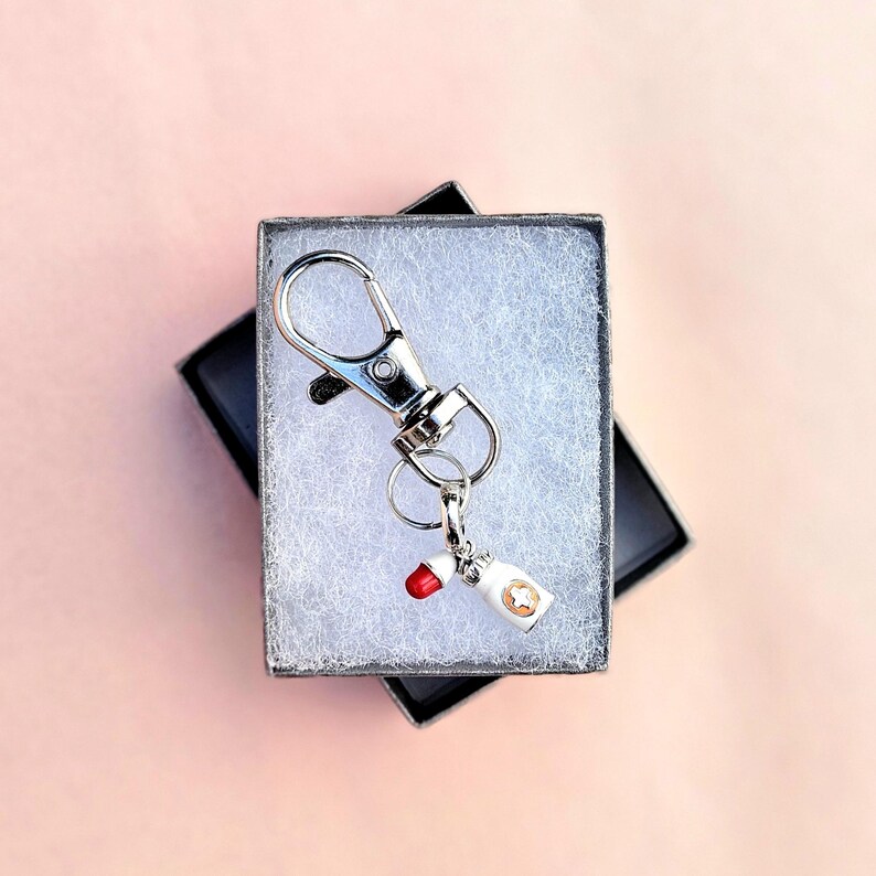 May include: A silver keychain with a lobster clasp and a small charm shaped like a white pill bottle with a red pill and a gold star. The keychain is presented in a small black gift box with a pink background.
