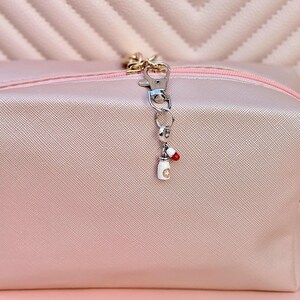 May include: A silver-tone charm with a white pill bottle and a red and white capsule attached to a clasp. The charm hangs from a pink cosmetic bag with a gold zipper.