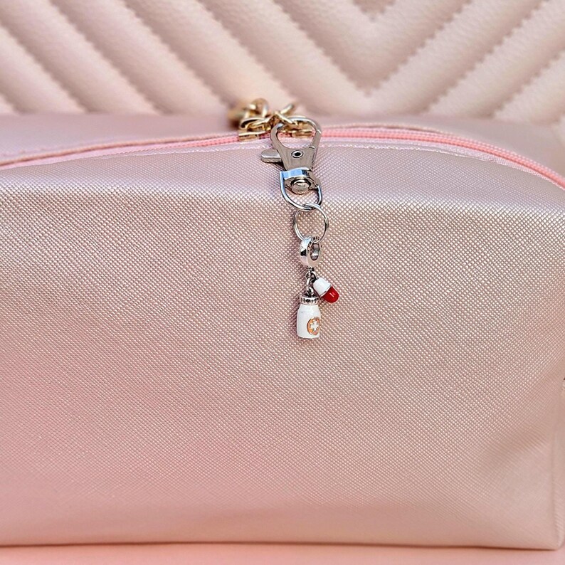 May include: A silver-tone charm with a white pill bottle and a red and white capsule attached to a clasp. The charm hangs from a pink cosmetic bag with a gold zipper.
