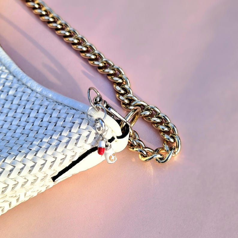 May include: A white woven handbag with a black stripe and a gold chain strap. A silver charm with a red and white pill design hangs from the bag's clasp. The background is a solid pink.