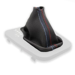 Könnte beinhalten: Schwarzer Lederschaltknauf mit roten und blauen Nähten. Die Nähte verlaufen als vertikaler Streifen in der Mitte des Schaltsacks. Der Schaltsack ist für den Schalthebel eines Fahrzeugs konzipiert und steht auf einer hellgrauen Basis.