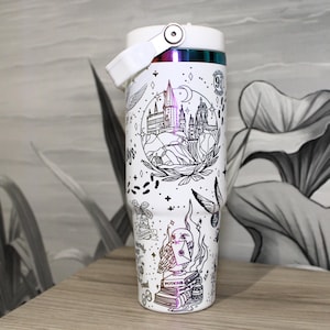 Puede incluir: Vaso térmico blanco con una ilustración en blanco y negro de un castillo, un búho y otros gráficos. El vaso tiene un asa y una tapa blancas con un acento de arcoíris. El diseño incluye el número 91.