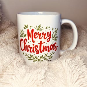 以下が含まれることがあります： 緑と金の植物柄が描かれた白いセラミック製のマグカップ。赤い文字で「メリークリスマス」と書かれている。