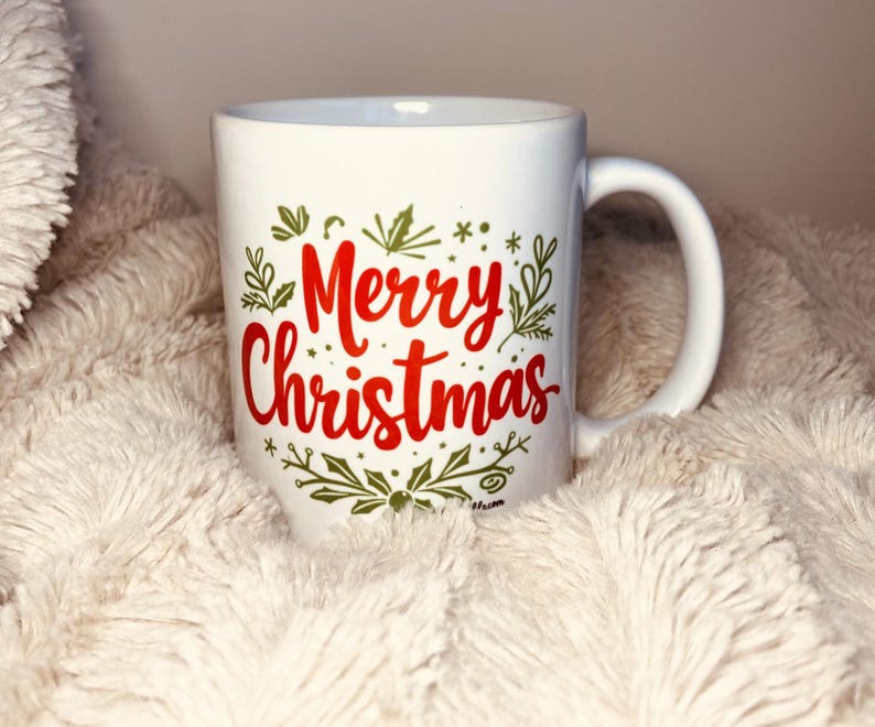 以下が含まれることがあります： 緑と金の植物柄が描かれた白いセラミック製のマグカップ。赤い文字で「メリークリスマス」と書かれている。