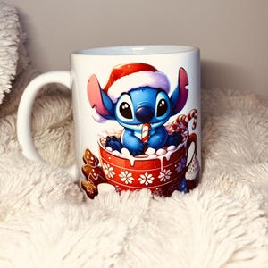 以下が含まれることがあります： 赤と白のサンタ帽をかぶった青と白の漫画のキャラクターが描かれた白いセラミックマグカップ。キャラクターはキャンディーケーンを持って、白いマシュマロでいっぱいの赤いマグカップに座っています。マグカップには赤と白の雪の結晶模様があります。