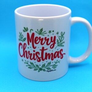 以下が含まれることがあります： 緑の葉で囲まれた赤い「メリークリスマス」のメッセージが書かれた白いセラミックマグカップ