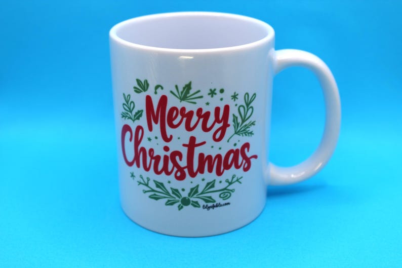 以下が含まれることがあります： 緑の葉で囲まれた赤い「メリークリスマス」のメッセージが書かれた白いセラミックマグカップ