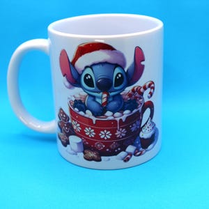 以下が含まれることがあります： 赤いサンタの帽子をかぶった青と白の漫画のキャラクターが描かれた白いセラミック製のマグカップ。キャラクターはキャンディーケーンを持って、マシュマロでいっぱいの赤いマグカップに座っています。マグカップの周りには、ジンジャーブレッドクッキー、キャンディーケーン、カップケーキなど、他のクリスマスのおやつがあります。