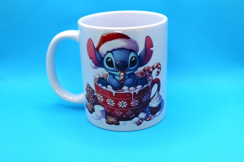 以下が含まれることがあります： 赤いサンタの帽子をかぶった青と白の漫画のキャラクターが描かれた白いセラミック製のマグカップ。キャラクターはキャンディーケーンを持って、マシュマロでいっぱいの赤いマグカップに座っています。マグカップの周りには、ジンジャーブレッドクッキー、キャンディーケーン、カップケーキなど、他のクリスマスのおやつがあります。