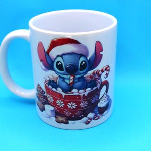 以下が含まれることがあります： サンタの帽子をかぶり、キャンディーケーンを持っているリロ＆スティッチのスティッチが描かれた、白いセラミック製のマグカップ。スティッチは、ホットチョコレートとマシュマロでいっぱいの赤いマグカップに座っています。