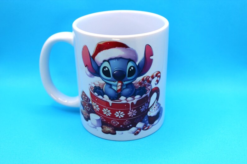 以下が含まれることがあります： サンタの帽子をかぶり、キャンディーケーンを持っているリロ＆スティッチのスティッチが描かれた、白いセラミック製のマグカップ。スティッチは、ホットチョコレートとマシュマロでいっぱいの赤いマグカップに座っています。