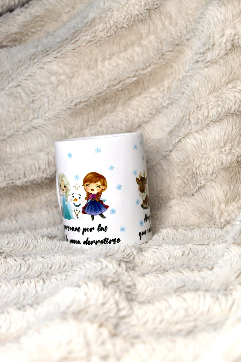 Puede incluir: Taza de cer&aacute;mica blanca con un dise&ntilde;o que presenta personajes de dibujos animados de la pel&iacute;cula Frozen. La taza tiene copos de nieve azules y el texto "Hay personas por las que vale la pena derretirse".
