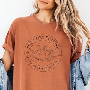 May include: A burnt orange t-shirt with a circular graphic that reads "THE COZY PUMPKIN" around a pumpkin illustration. The text "EST 2020" and "FARM FRESH PUMPKINS" are also included. The shirt is a relaxed fit.