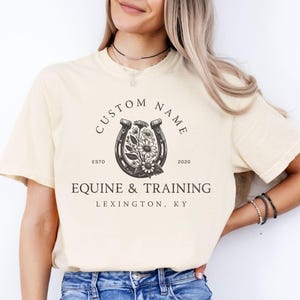 May include: Ivory-colored t-shirt featuring a horseshoe design with floral elements and the text "CUSTOM NAME" above and "EQUINE & TRAINING LEXINGTON, KY" below. The shirt also has the text "ESTD 2020".