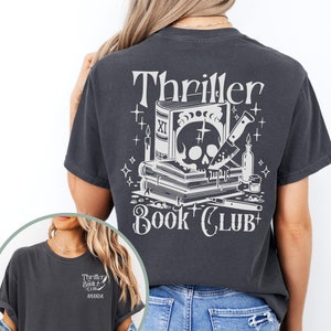 May include: Dark gray t-shirt with a white graphic on the back that reads "Thriller Book Club." The graphic includes a skull, books, a knife, and other spooky elements. The front of the shirt has a smaller graphic with the same text.