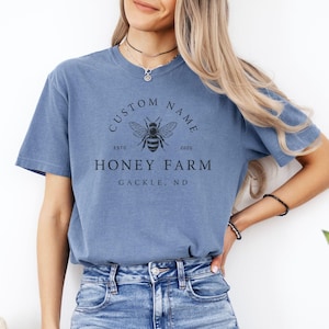 May include: A blue t-shirt with a vintage-style logo featuring a bee and the text "CUSTOM NAME ESTD 2000 HONEY FARM GACKLE, ND".