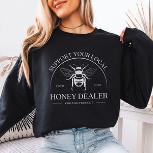 May include: Black sweatshirt with a white graphic featuring a bee and the text "SUPPORT YOUR LOCAL HONEY DEALER ORGANIC PRODUCT". The design includes the establishment year 2020. The sweatshirt is a solid black color.