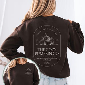 May include: Dark chocolate brown sweatshirt with a white graphic on the back featuring pumpkins and the text "THE COZY PUMPKIN CO." The front of the sweatshirt is visible with the same logo. The sweatshirt is a fall-themed product.