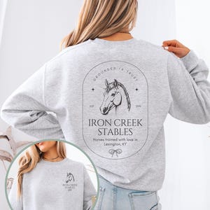 May include: Light gray sweatshirt with the text "IRON CREEK STABLES" and a horse head logo. The back of the sweatshirt features the same logo and text, with the words "Grounded in Trust" above the horse head. The front of the sweatshirt is also visible.