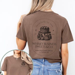 May include: Espresso-colored t-shirt featuring a graphic of a cake and other baked goods, with the text "WHISKY BUSINESS SWEETS & CO." and "sweet treats baked with love in Portland, ME." The shirt also has the text "FRESH FROM THE OVEN" and "EST. 2010."