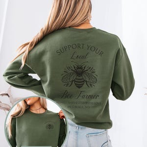 May include: Olive green sweatshirt with the text "SUPPORT YOUR Local Bee Farmer" and a bee graphic. The text "HONEY HARVESTED WITH LOVE IN GACKLE, ND" is below the bee graphic. A smaller image of the sweatshirt front is in the lower left.