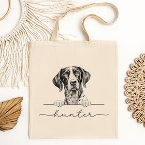 May include: A beige tote bag featuring a black and white dog illustration. The dog is peering over a horizontal line, with the name "hunter" written in a cursive font below. The bag has beige handles.