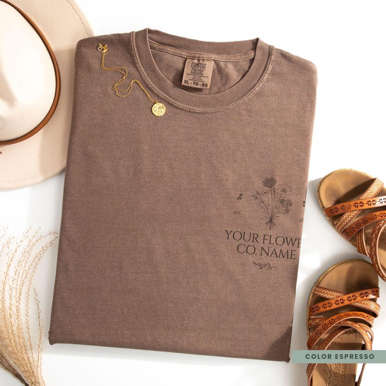 May include: A brown t-shirt with a floral design and the text "YOUR FLOWE CO. NAME". A gold necklace, a white hat, and brown sandals are arranged around the shirt. The shirt is labeled "COLOR ESPRESSO".
