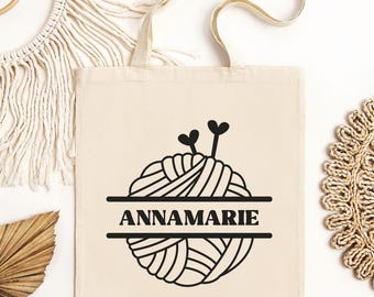 May include: Cream-colored tote bag featuring a black graphic of a yarn ball with knitting needles and the name "ANNAMARIE" in a bold font. The bag has a simple design with two handles.