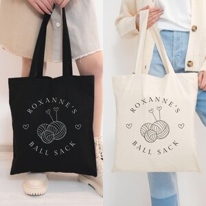 May include: Two tote bags with the text "Roxanne's Ball Sack" and a graphic of yarn balls and knitting needles. One bag is black and the other is natural canvas.