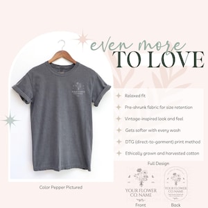 May include: A gray t-shirt with rolled-up sleeves hangs on a wooden hanger. The shirt features a small floral design and text on the front. The text "even more TO LOVE" is at the top. The color of the shirt is "Pepper".