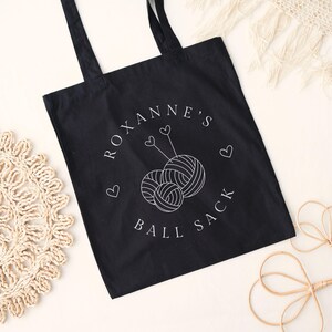 May include: Black tote bag with white text that reads "Roxanne's Ball Sack" and a graphic of two yarn balls with knitting needles.