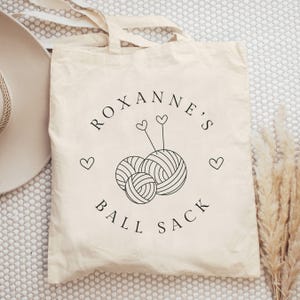 May include: A white canvas tote bag with black text that reads "Roxanne's Ball Sack" and a graphic of two yarn balls with needles.