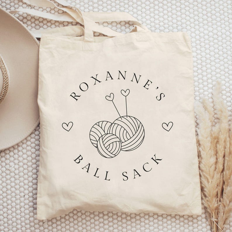 May include: A white canvas tote bag with black text that reads "Roxanne's Ball Sack" and a graphic of two yarn balls with needles.