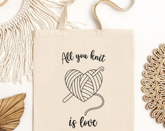 May include: A white canvas tote bag with black text that reads "All you knit is love". The text is accompanied by a black and white illustration of a heart-shaped ball of yarn with a crochet hook.