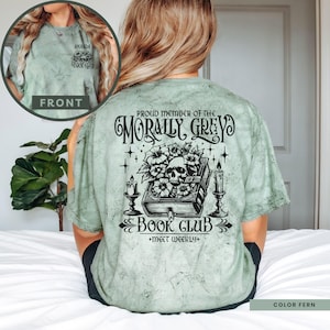 May include: A green t-shirt with a black and white graphic design featuring a skull, flowers, and a book. The text on the shirt reads "Proud member of the Morally Grey Book Club + Meet Weekly+".